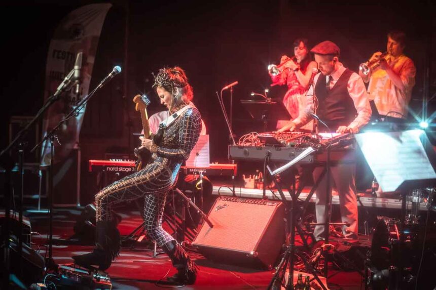 Po lewej kobieta w obcisłym błyszczącym stroju gra na gitarze, po prawej reszta zespołu