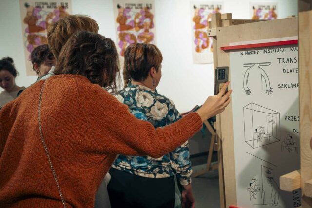 ludzie tyłem oglądają rysunki na drewnianych stelażach