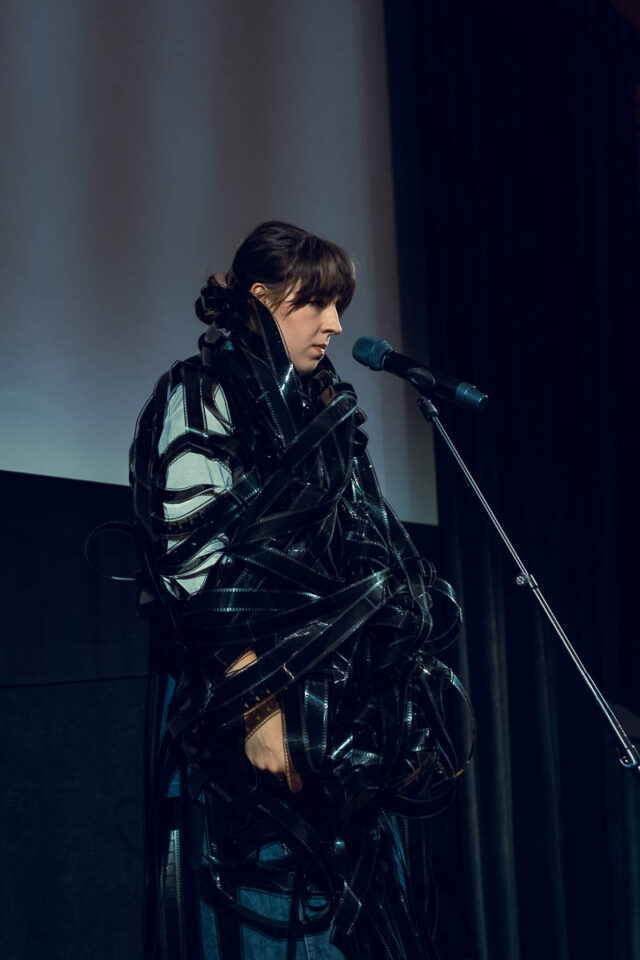 Młoda kobieta przy mikrofonie owinięta czarną taśmą filmową