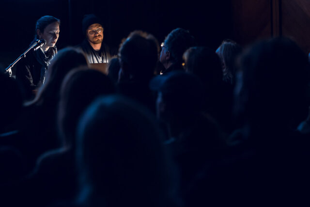 na pierwszym planie publiczność w ciemności, w tle oświetlone dwie postaci kobiety i mężczyzny