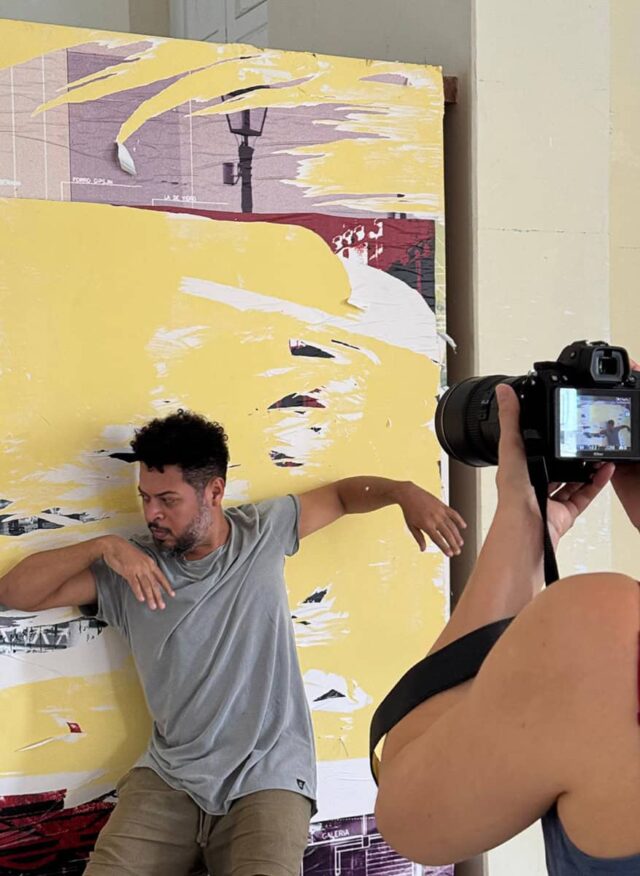 Na pierwszym planie dłoń z aparatem, na drugim mężczyzna w tanecznej pozie na tle obrazu