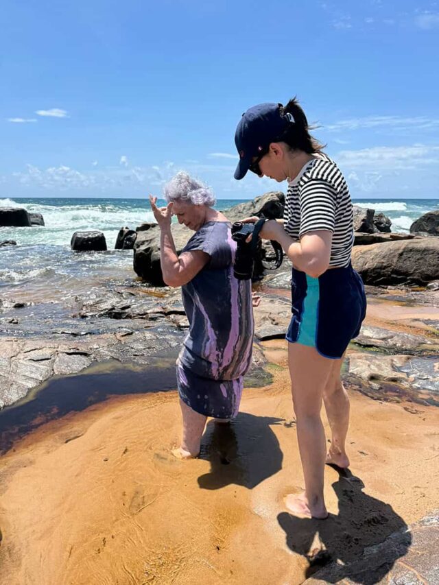 Starsza kobieta tańczy na tle morza, na pierwszym planie druga z aparatem foto