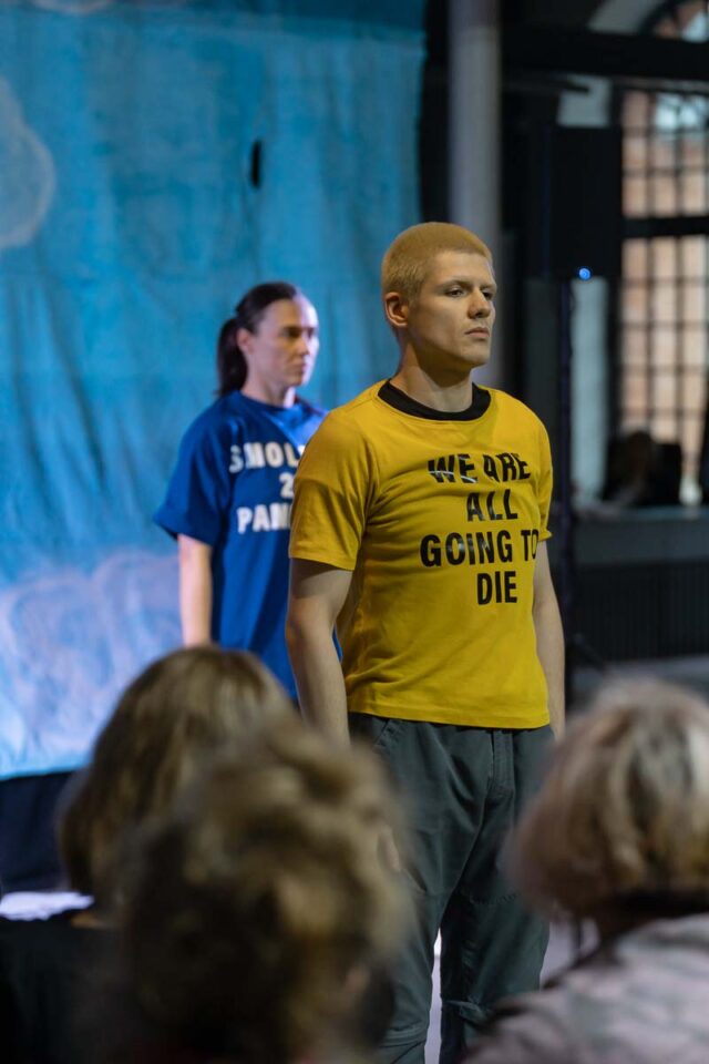 mężczyzna o jasnych włosach w żółtej koszulce, w tle kobieta w niebieskiej