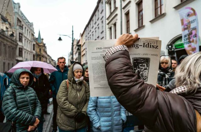 Kobieta stojąca tyłem pokazująca gazetę Jung Jidysz grupie ludzi