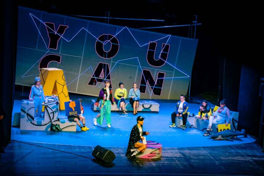 Aktorzy w kolorowych strojach na dużej scenie, w tle duży napis YOU CAN