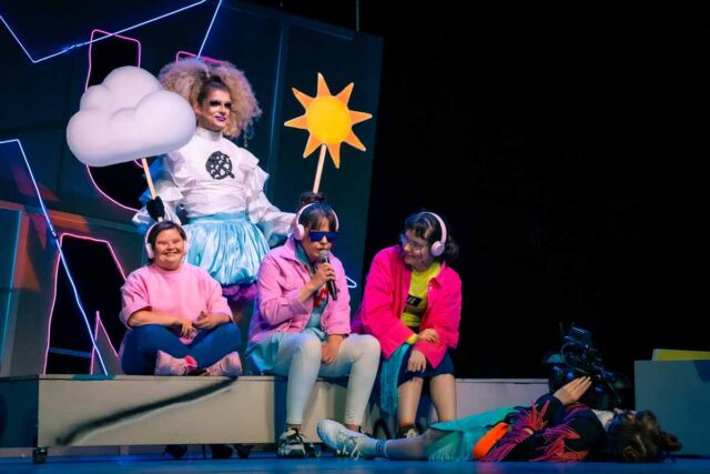 3 kobiety w różowych bluzach siedzą na podeście za nimi drag queen z kartonowym słońcem i chmurką w dłoniach a przed nimi na ziemi lezy osoba z kamerą