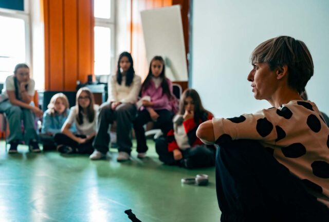 Kobieta o krótkich włosach - reżyserka - kuca na pierwszym planie, w tle grupa młodziezy