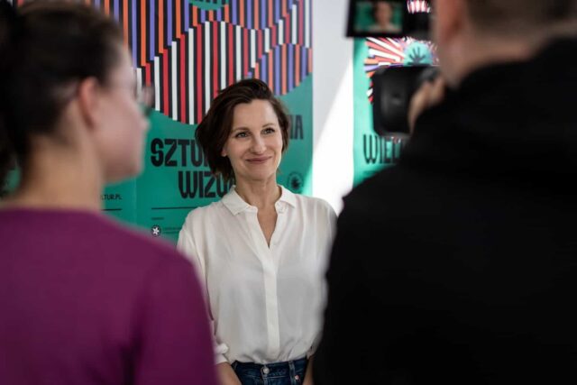 Kobieta w białej bluzce na tle plakatów, na pierwszym planie dwie postaci tyłem