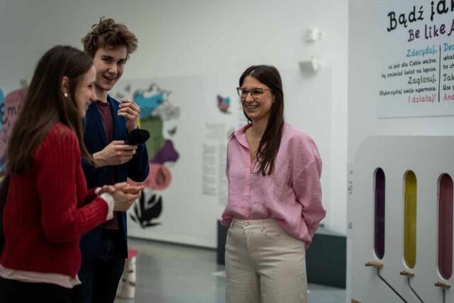 kobieta w różowej bluzce po prawej udziela wywiadu parze młodych ludzi