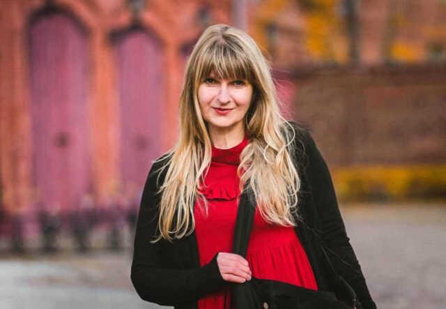 Kobieta w długich blond włosach, czerwonej bluzce i czarnym swetrze na tle starej architektury