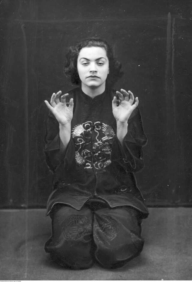 Czarno-białe archiwalne zdjęcie kobiety w ciemnym stroju z uniesionymi dłońmi