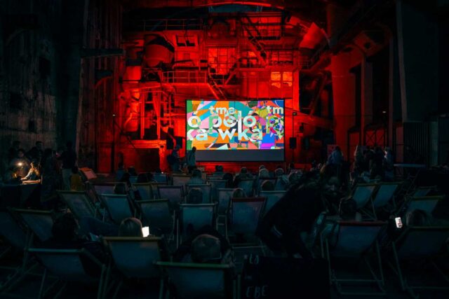 Ciemne, podświetlone na czerwono wnętrze fabryki. Na pierwszym planie ludzie na leżakach, w tle duży ekran.