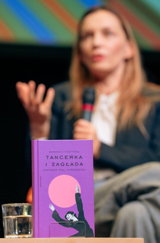 na pierwszym planie książka, w tle kobieta - blondynka z mikrofonem (rozmyta)