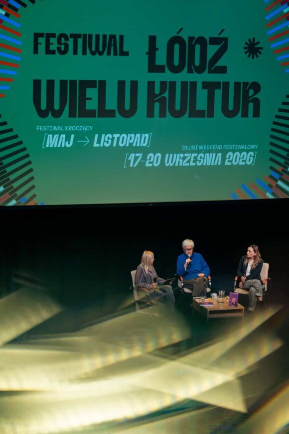 w oddali 3 osoby siedzą na krzesłach na scenie, w tle baner z napisem Festiwal Łódź Wielu Kultur. Na pierwszym tle świetliste maziaje