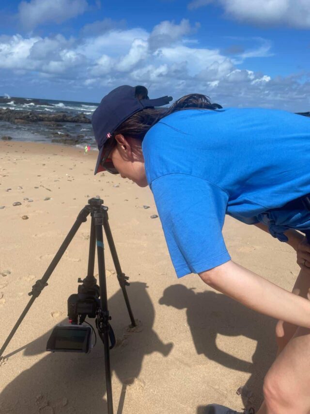 Kobieta w niebieskiej koszulce nachyla się nad statywem fotograficznym na plaży