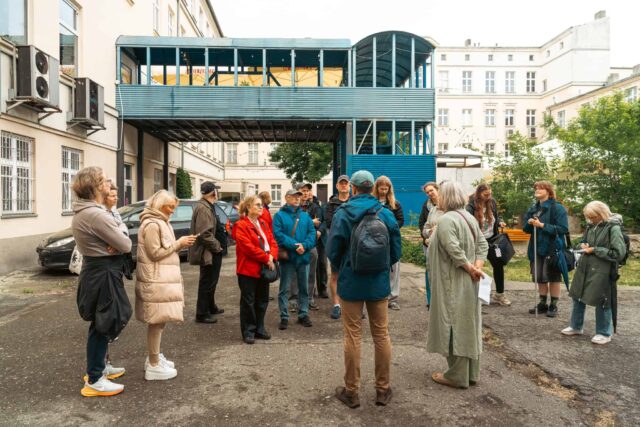 Grupa osób stojąca na podwórku, przewodnicy tyłem