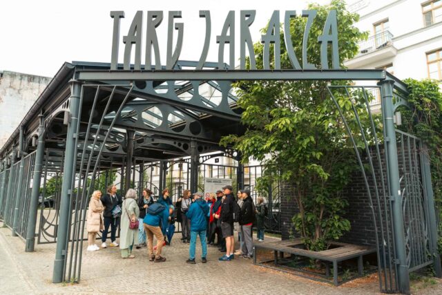 grupa osób stojąca pod konstrukcją metalową z napisem Targ Jaracza