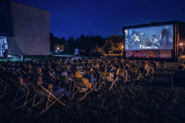 Na pierwszym planie ludzie siedzą na leżakach tyłem, w tle ekran na którym leci film