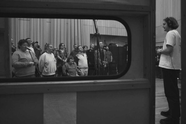 czarno białe zdjęcie z backstagu nagrywania płyty, po prawek męzczyzna w białym t-shircie, po lewej przez okno widać zespół