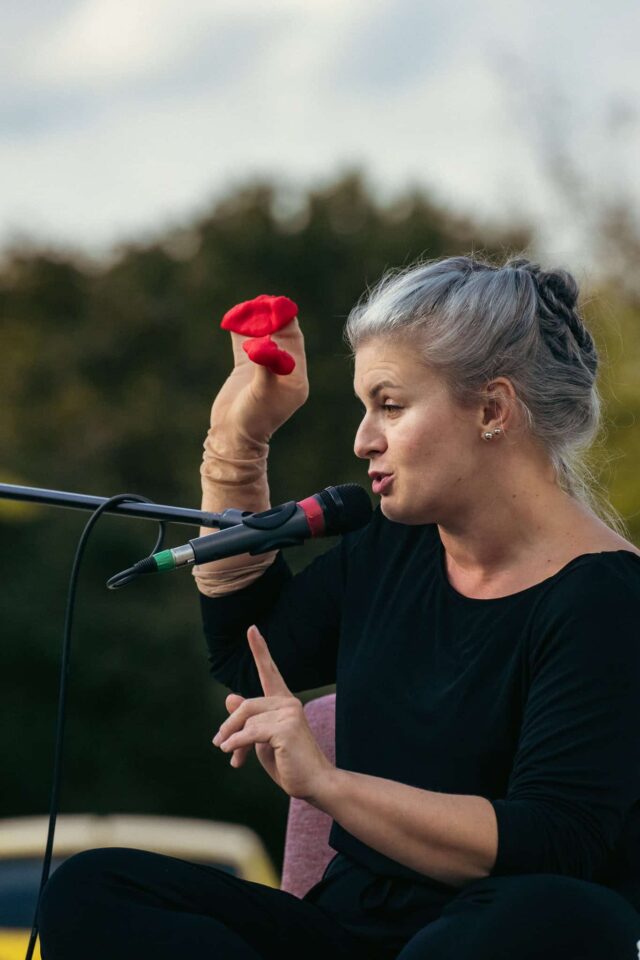 Kobieta przy mikrofonie, w dłoniach trzyma coś czerwonego