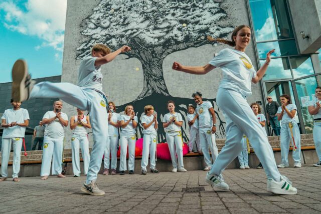 kilkanaście osób w białych strojach, dwie na pierwszym planie pokaz capoeiry