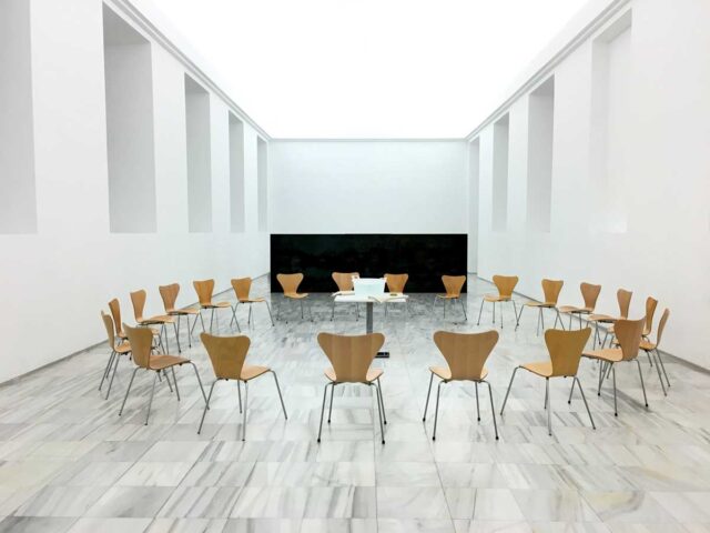 Jasna sala galerii z drewnianymi krzesłami ustawionymi w dużym kręgu wokół centralnego stołu.