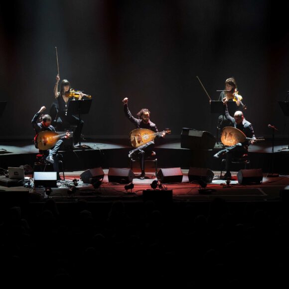 Trzech mężczyzn gra na instrumentach na scenie w ciemnej przestrzeni