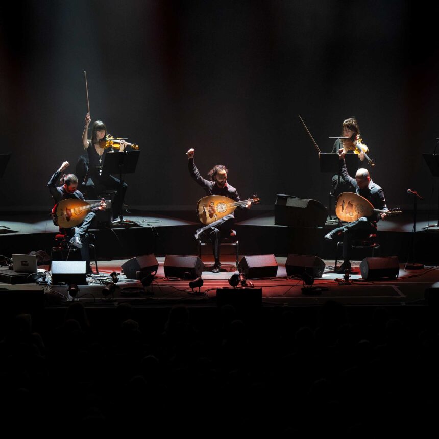 Trzech mężczyzn gra na instrumentach na scenie w ciemnej przestrzeni
