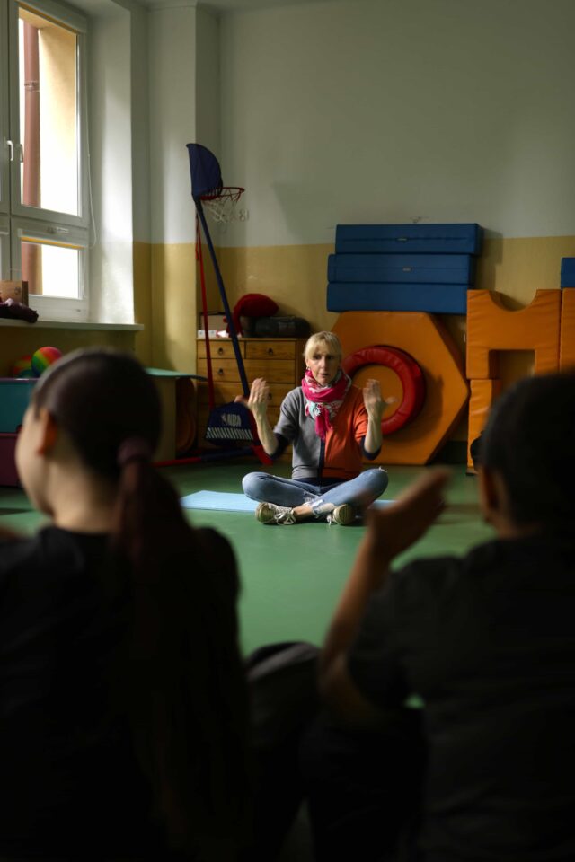Kobieta siedzi na podłodze, na pierwszym planie zarysy sylwetek dzieci